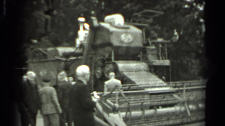 1963 Russia 8mm frame with men clustered near a combine harvester, the header and side tracks visible in a wooded outdoor setting.