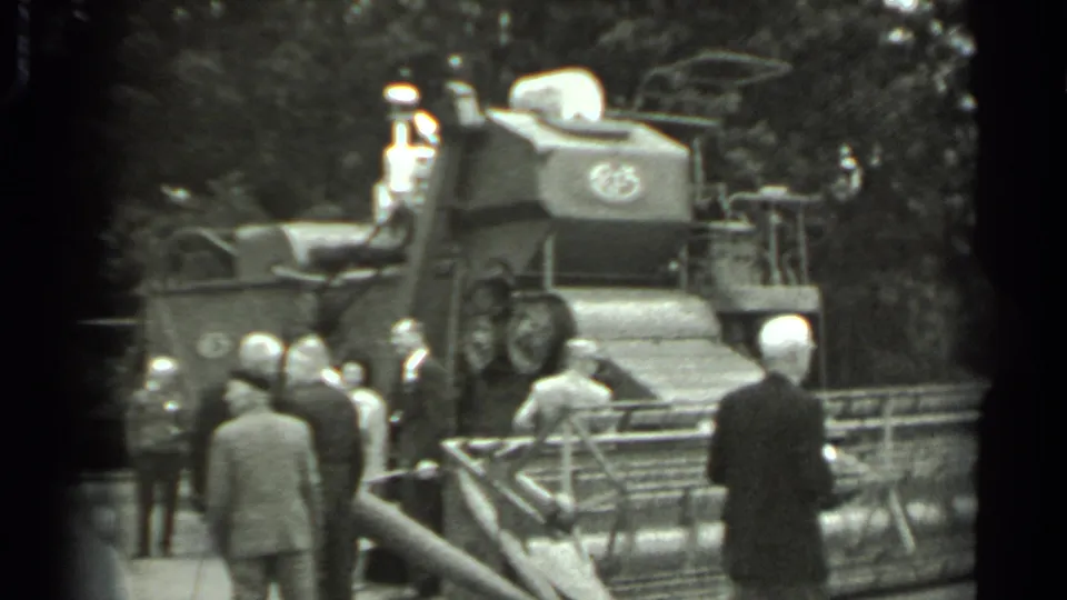 1963 Russia home-movie frame showing a group of men in formal attire near a large combine harvester with trees in the background.
