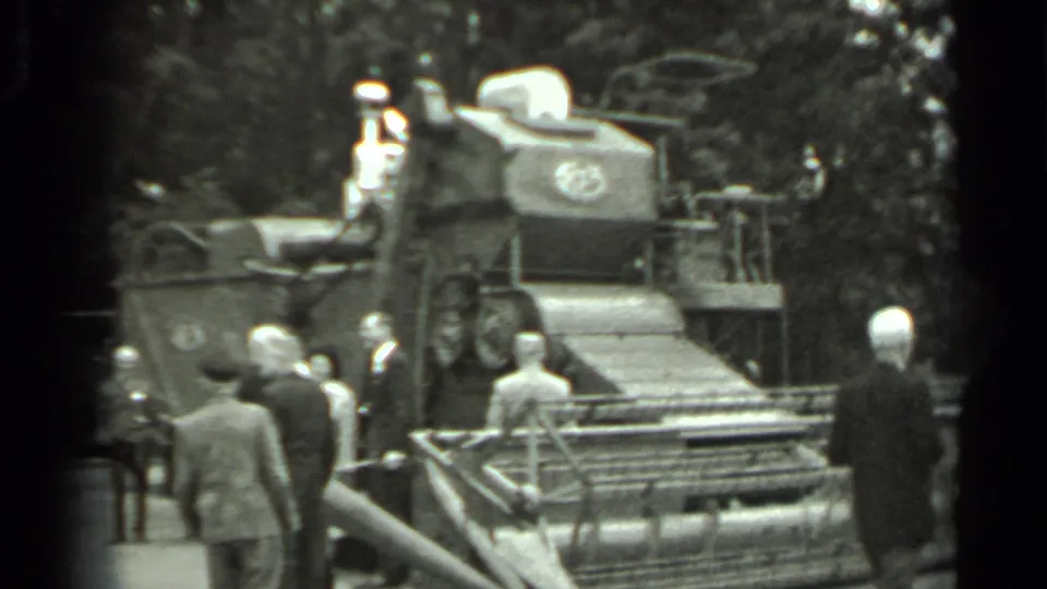 1963 Russia 8mm frame showing suited visitors viewing a large combine harvester with a front cutting mechanism in a tree-lined outdoor display.