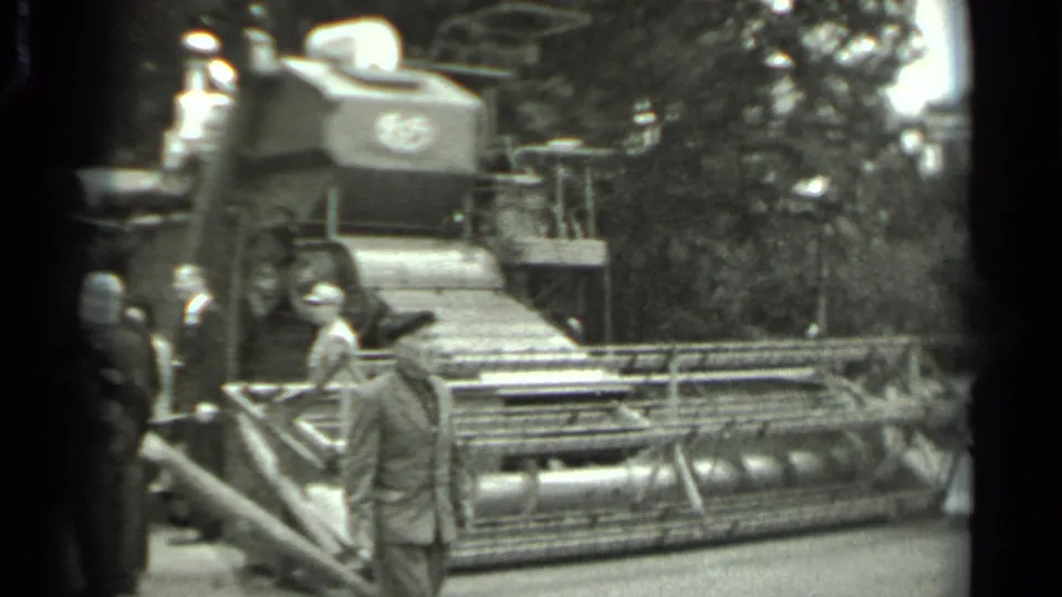 1963 Russia 8mm frame showing a man in period clothing passing a large combine harvester at a wooded outdoor machinery display.