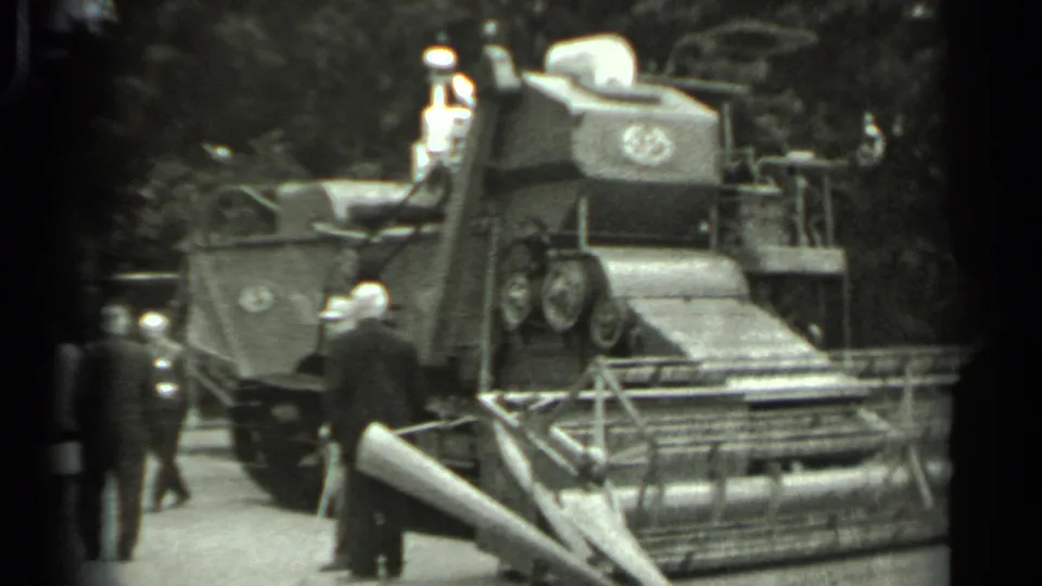 1963 Russia 8mm frame with men viewing the side of a tracked harvester, its reel partly visible in the foreground.