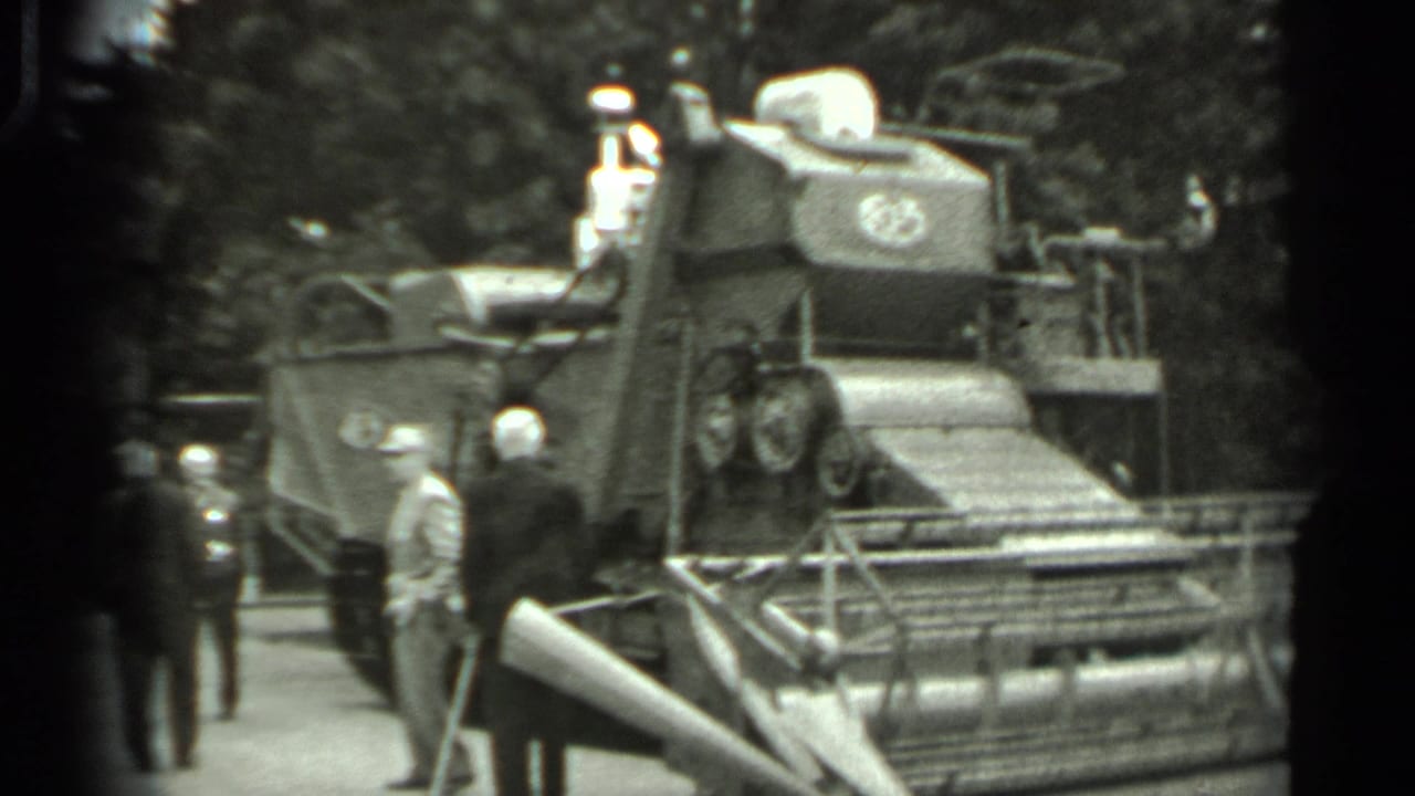 Men stand beside a large tracked agricultural machine with a cutting header.