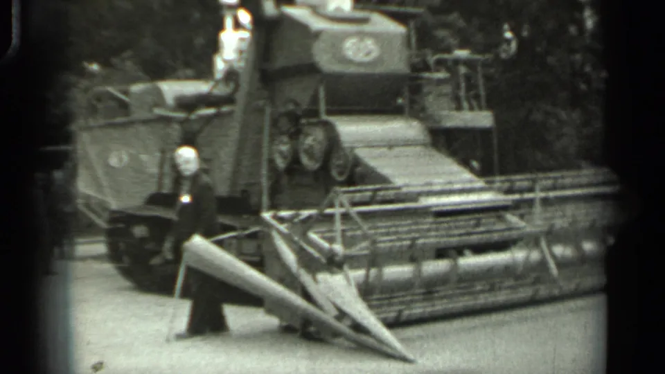 1963 Russia home-movie frame showing a suited visitor near the header of a large tracked agricultural machine.