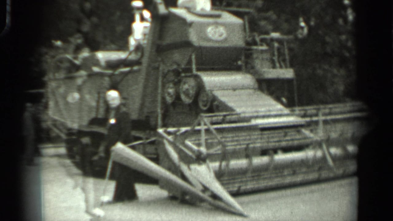 Man in a light hat stands beside a tracked agricultural machine with a front cutting header.