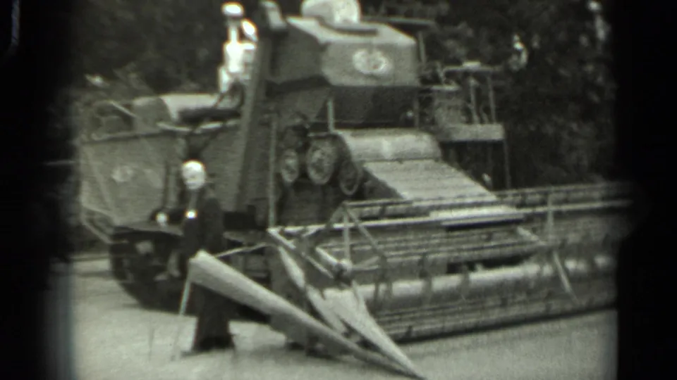 Black-and-white 8mm frame from Russia, 1963, showing the agricultural machine's tracks and header with a demonstrator nearby.