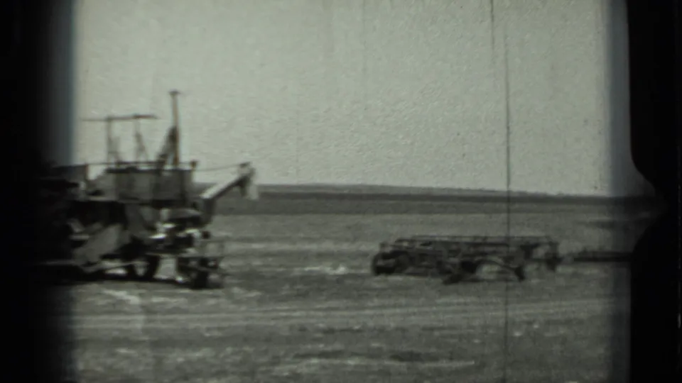 1963 Russia 8mm frame showing the edge of a parked vehicle and low field equipment on flat rural terrain.
