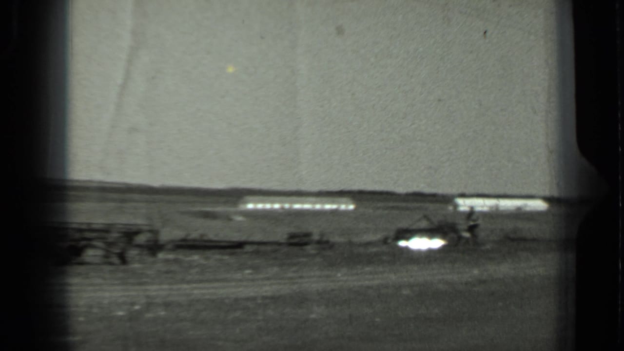 Black-and-white 1963 Russia home-movie frame of an open field, distant low structure, and small agricultural equipment in soft focus.
