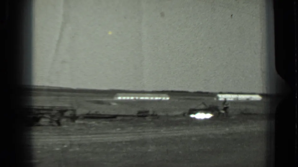 Black-and-white 1963 Russia home-movie frame of an open field, distant low structure, and small agricultural equipment in soft focus.