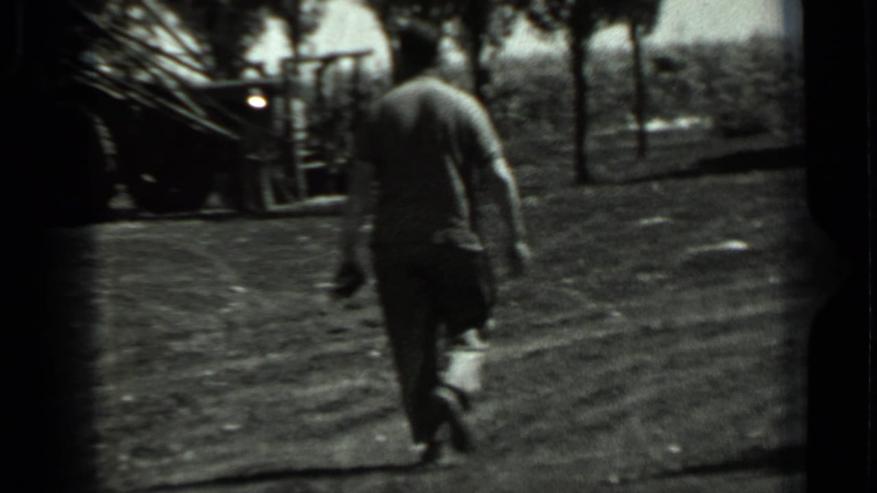 A man walks through a rural Soviet field toward machinery and trees in grainy black-and-white 8mm footage.