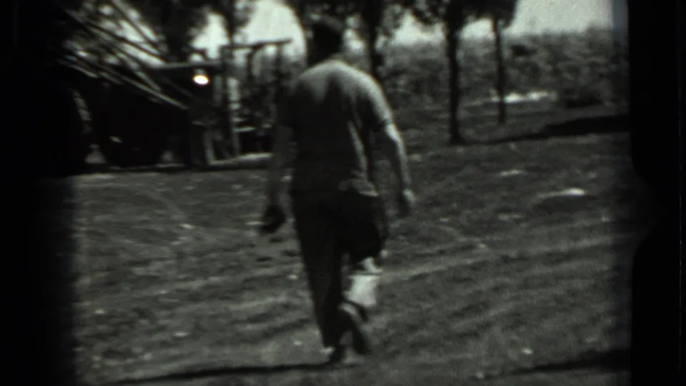 A man walks through a rural Soviet field toward machinery and trees in grainy black-and-white 8mm footage.