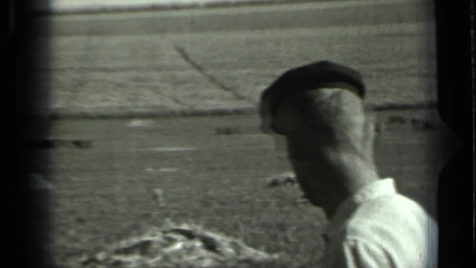 Black-and-white 8mm frame of a cap-wearing adult man in profile near a rural Russian field with distant cattle behind him.