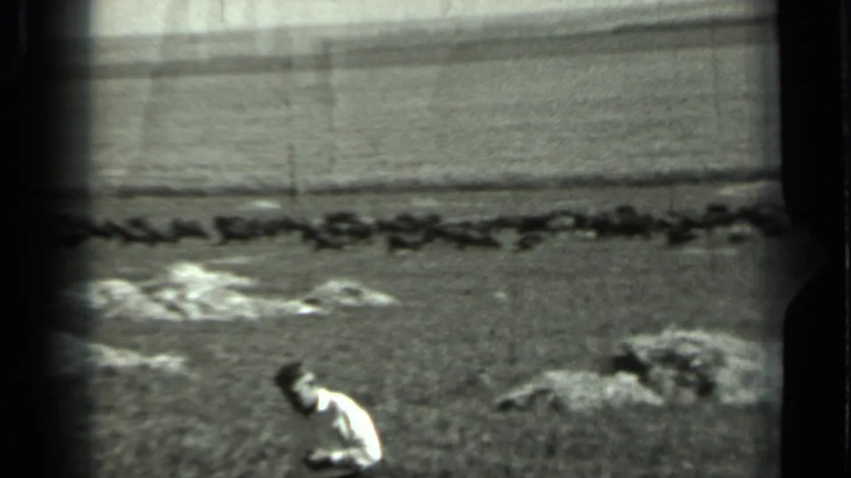 1963 Russia home-movie frame showing a man in light clothing walking across the foreground of an open field with rocks and a distant horizon.