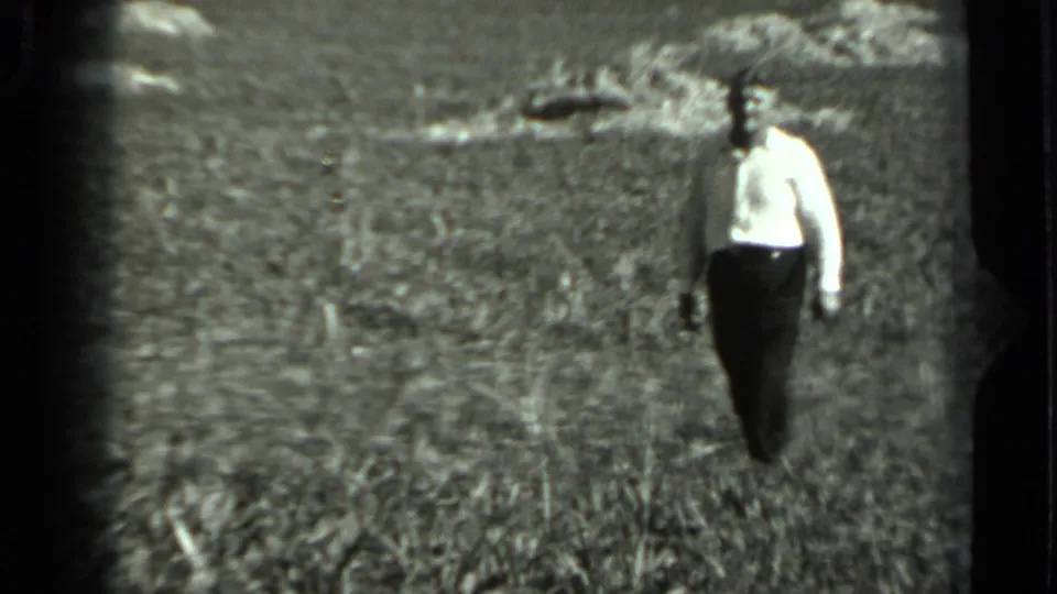 In 1963 Russia home-movie footage, a man in a light shirt walks through tall grass with distant animals across the field.