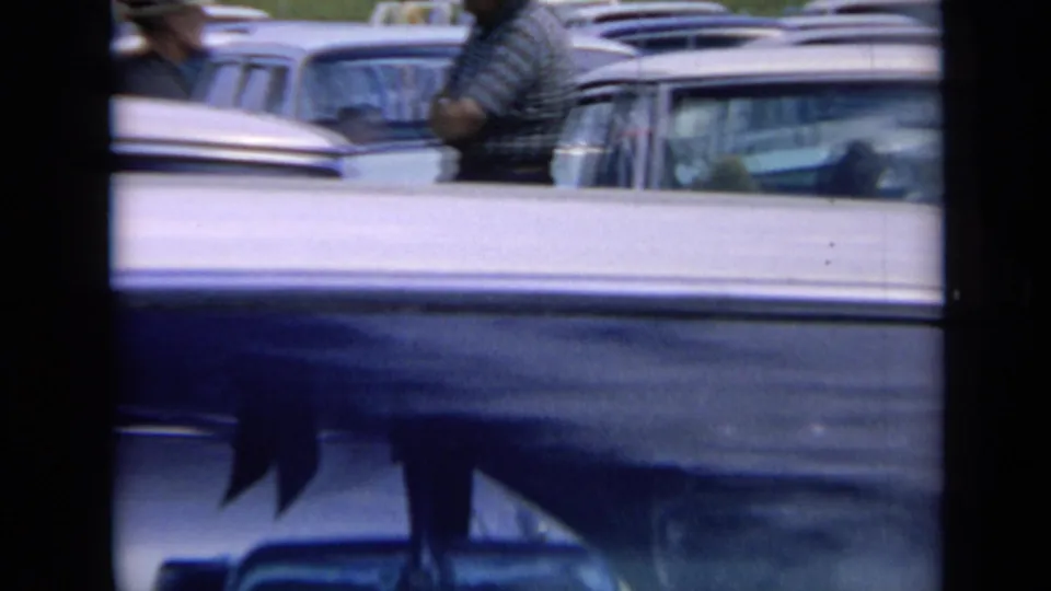 1964 Kansas home-movie frame showing parked mid-century cars and partial adult figures near the cattle area.