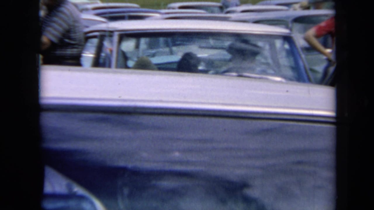 1964 Kansas home-movie frame showing closely packed sedans and wagons in a traffic jam, with people standing among the cars in bright daylight.