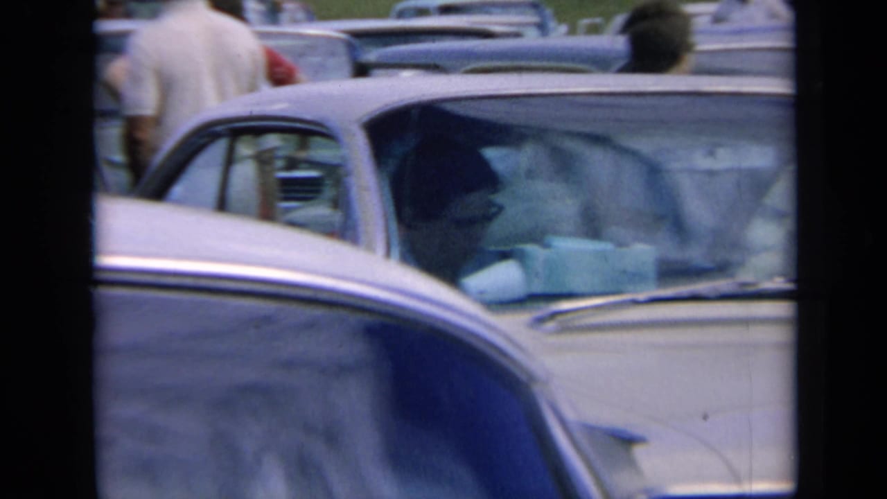 1964 Kansas traffic-jam frame with overlapping car roofs, a pale sedan at right, a person seated inside, and another person standing between vehicles.