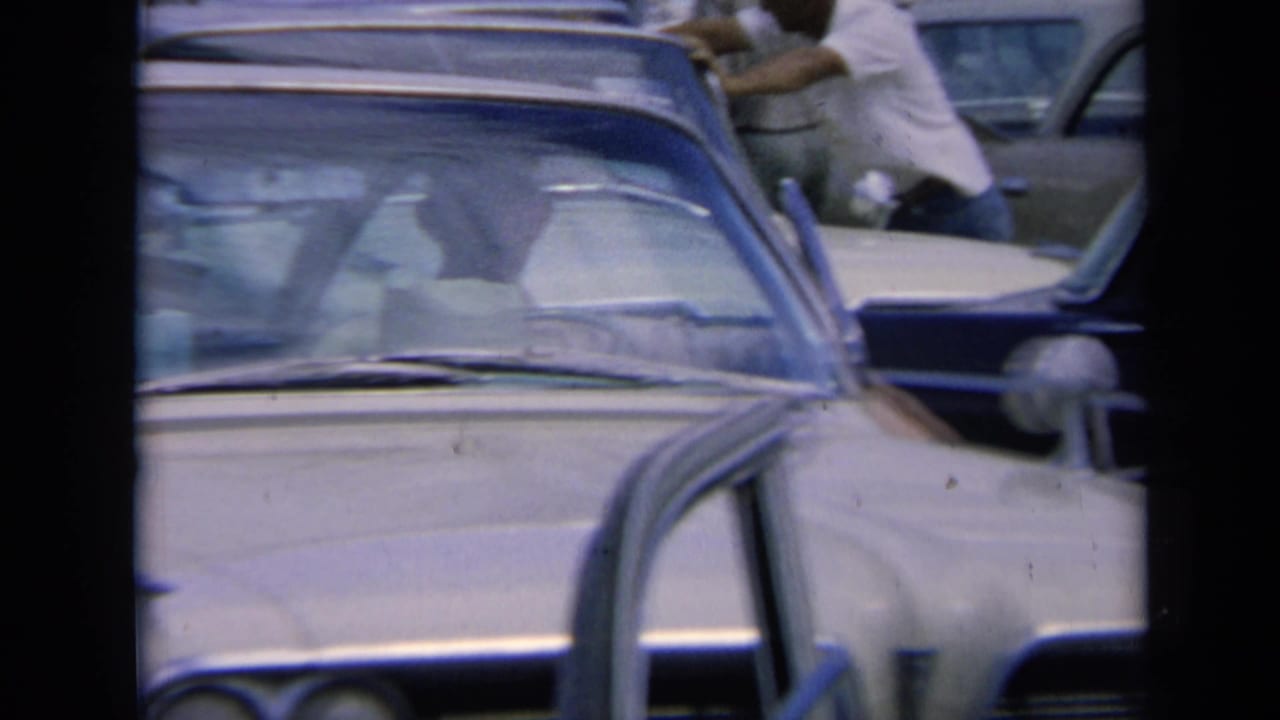 1964 Kansas home-movie frame showing the front of a light-colored sedan, surrounding cars, and partially visible people moving or standing in the traffic jam.