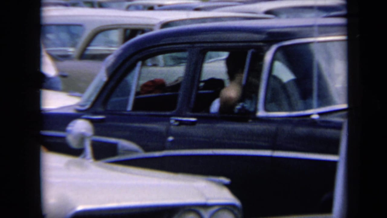 1964 Kansas frame with a dark station wagon in the foreground, a light-colored car nose below it, and rows of vehicles packed together in a daylight traffic jam.