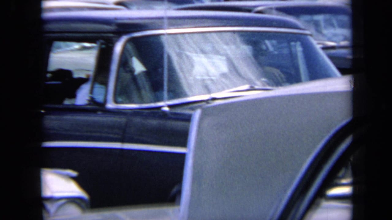 1964 Kansas home-movie frame showing tightly packed classic American cars, windshields, and chrome trim viewed through dark foreground bars or window edges.