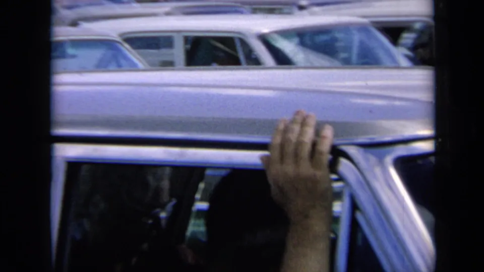 1964 Kansas home-movie frame showing a raised hand beside or above a car roof, with tightly grouped mid-century vehicles filling the background.