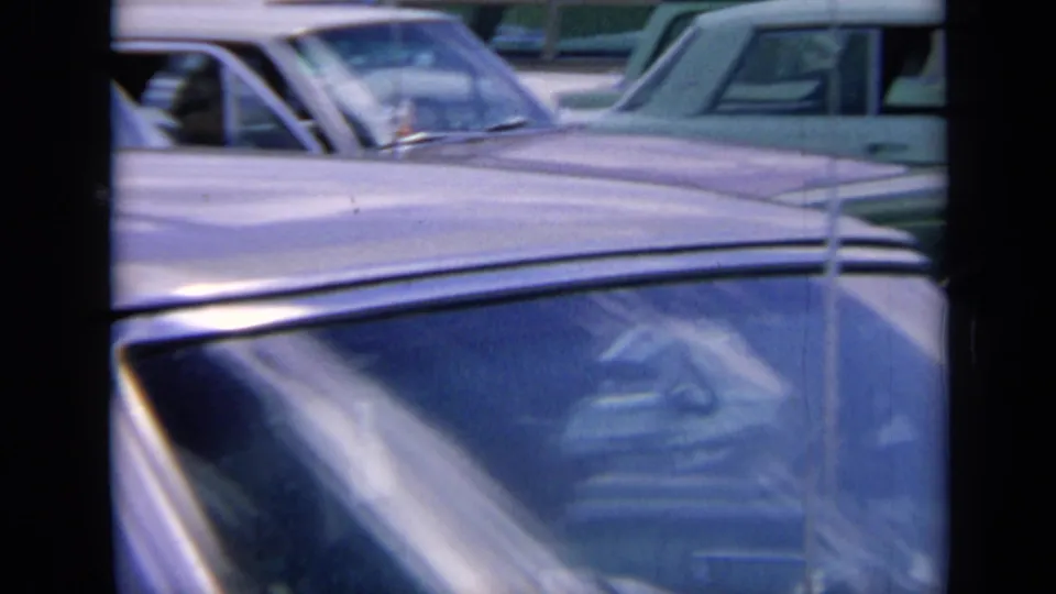 1964 Kansas traffic frame showing a close foreground car windshield, chrome roofline, window reflections, and additional stalled cars behind it.