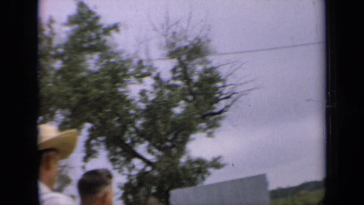 1964 Kansas 8mm frame with two partly visible adults near a farm setting, one in a straw hat, with trees and power lines behind them.