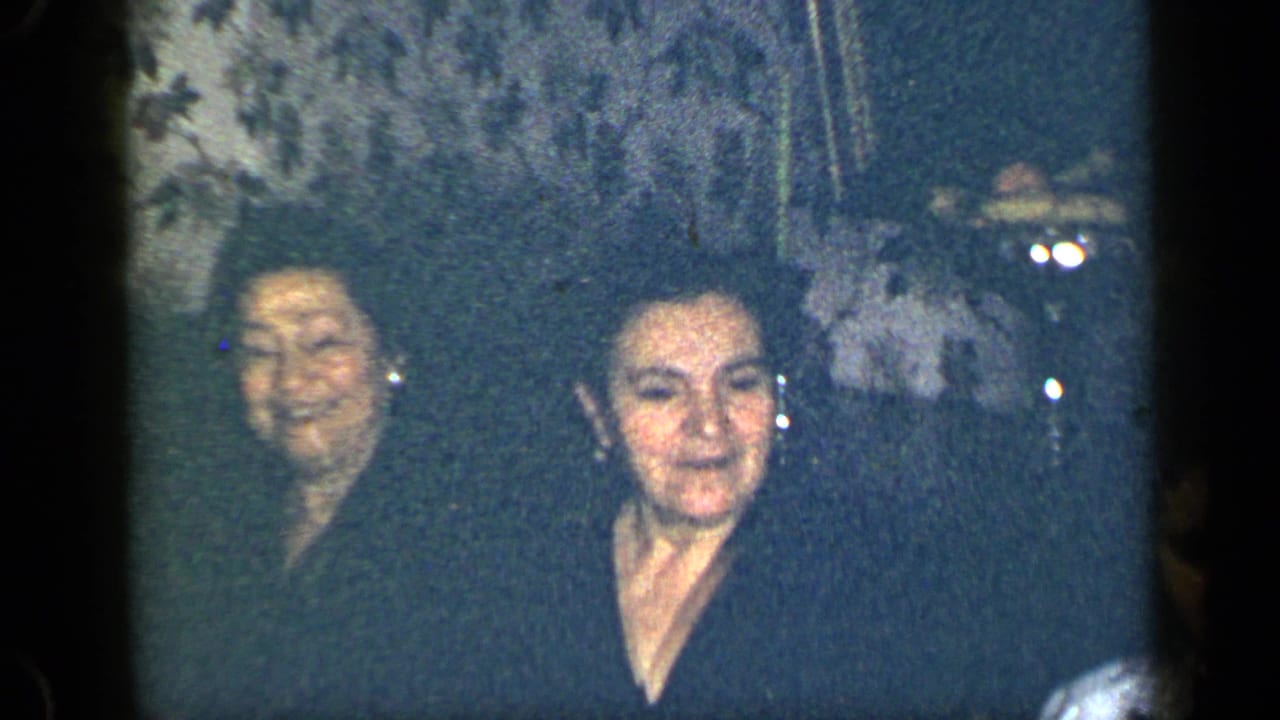 Two older women in dark clothing sit or stand indoors against patterned wallpaper.