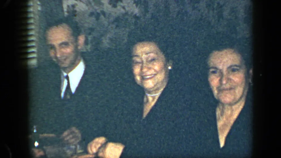In a 1948 New York home-movie interior, a suited man stands beside two women near patterned wallpaper and shutters.