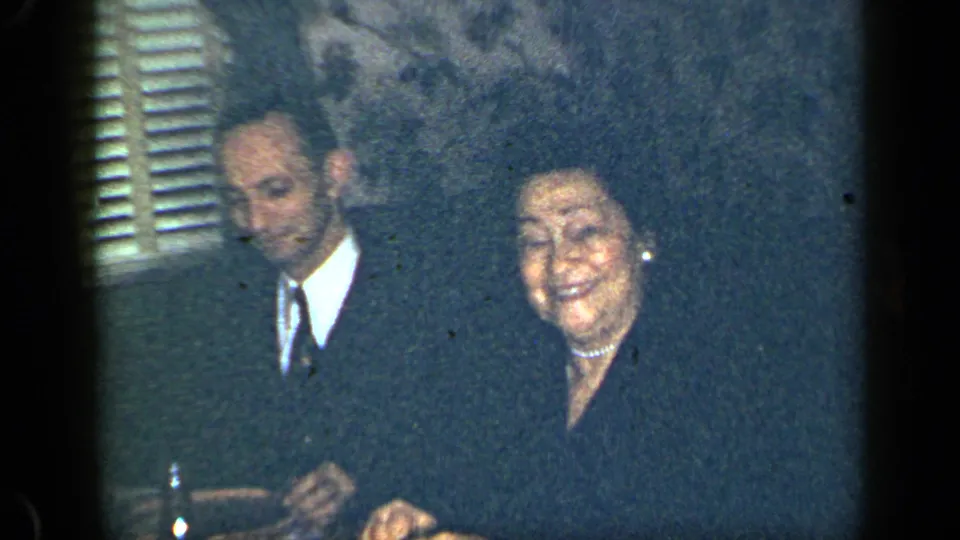 A suited man and smiling older woman appear in a dim 1948 New York private interior with shutters and patterned wallpaper.