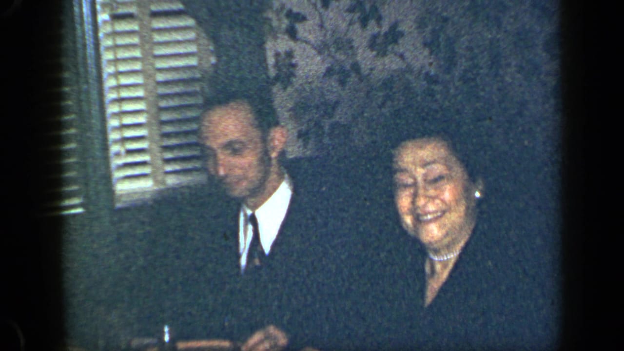 Suited man and smiling woman seated at a table near shuttered windows.