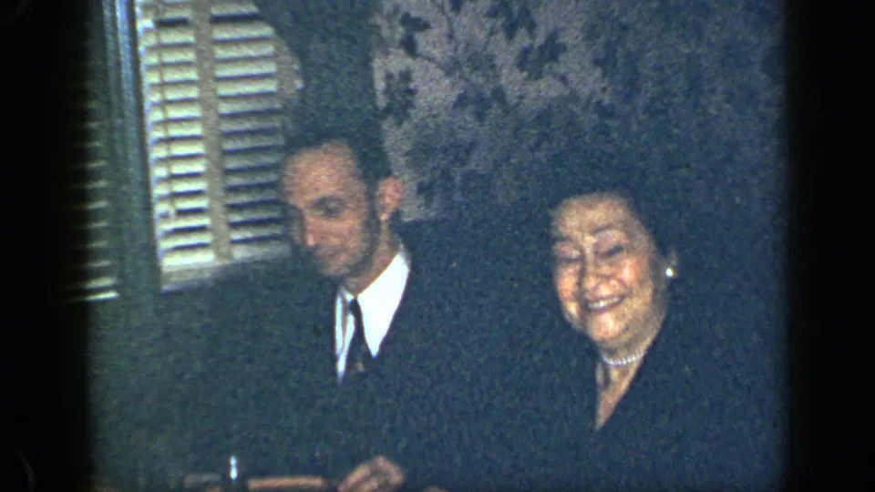1948 New York home-movie frame showing a suited man and smiling woman seated indoors by shuttered windows.