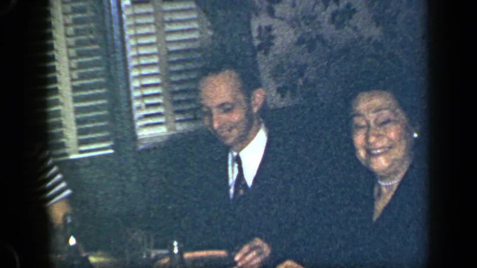 The man in a suit and the woman in a dark dress remain seated near the table in the New York interior.