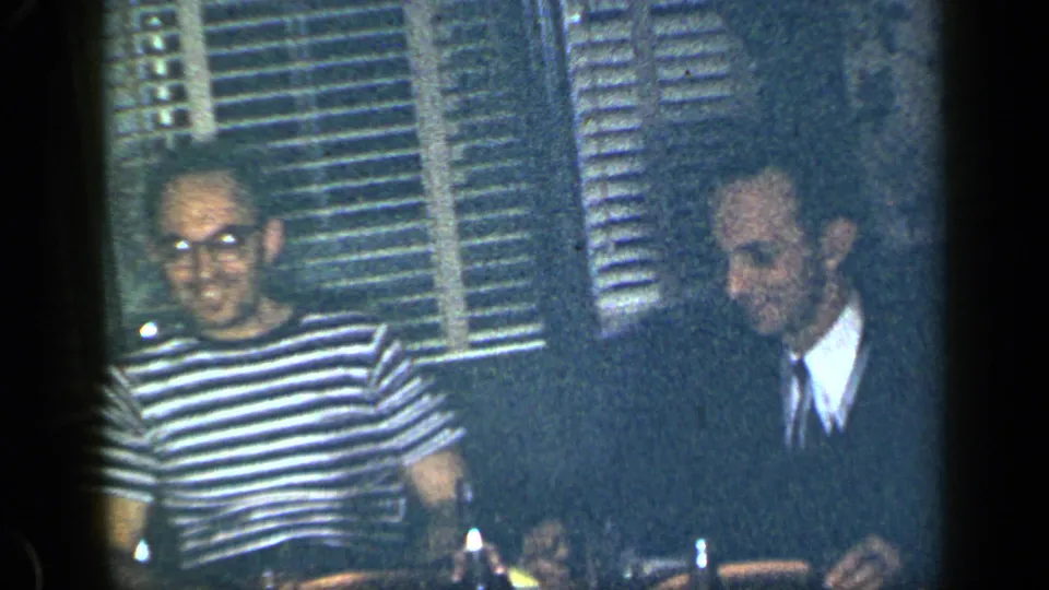 A man in a striped shirt and glasses sits near a suited man at a table in the 1948 New York home movie.