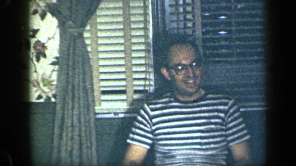 The striped-shirted man with glasses sits by the shuttered window in a private New York interior.