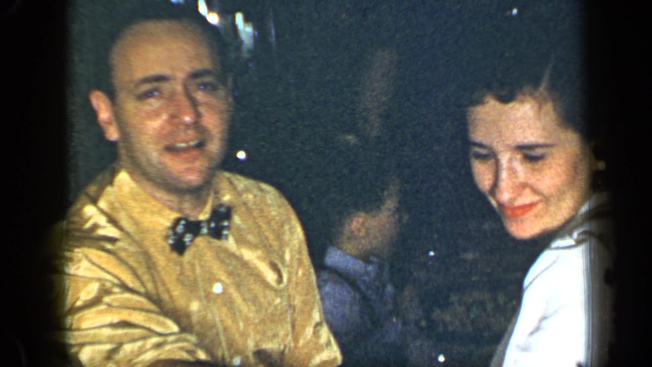 Man in gold shirt and bow tie sits beside a woman in white at a dim dinner table.