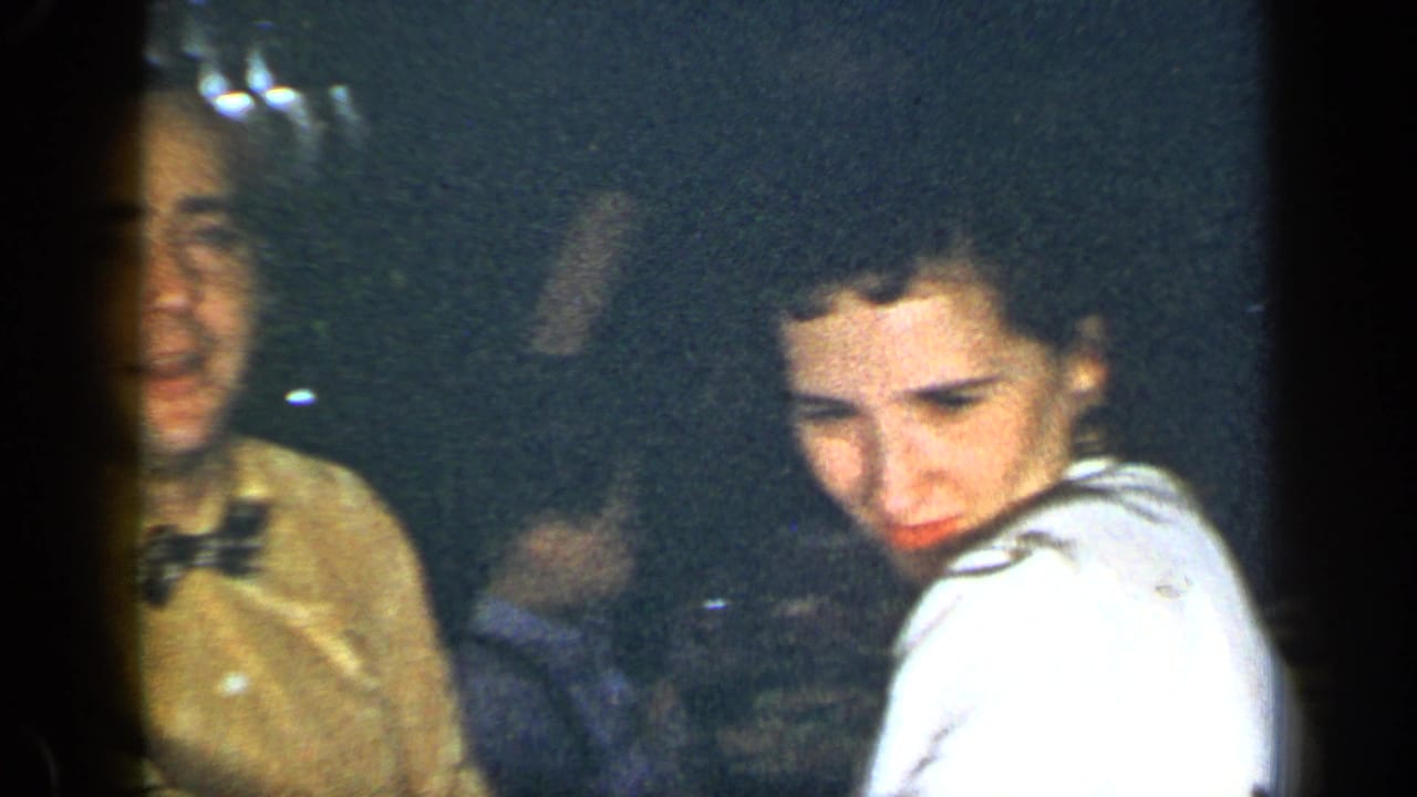 Two dinner guests sit in dim light, with a woman in white looking down near the table.