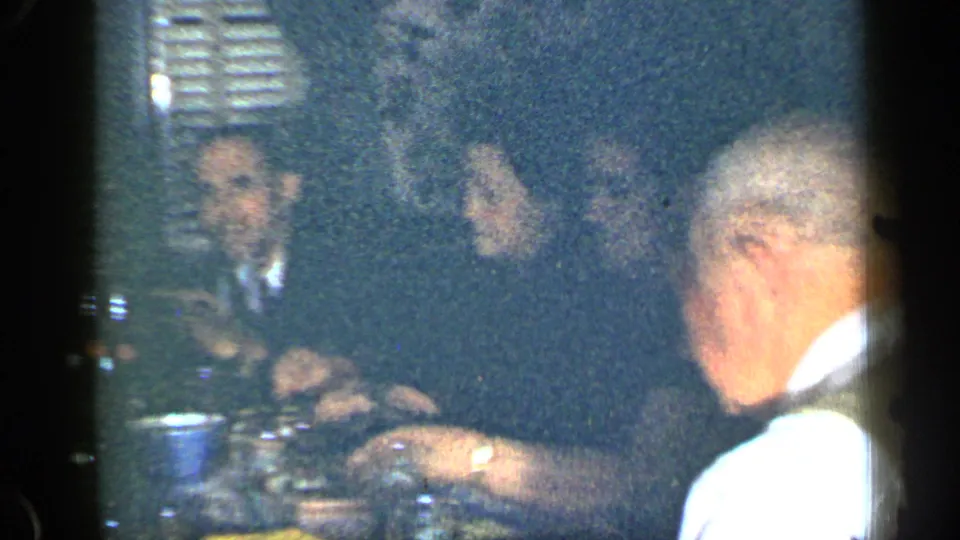 Grainy 1948 New York home-movie frame of adults seated around a dining table with cups, glasses, plates, and food.