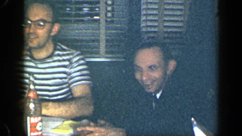 1948 New York home-movie frame of two seated men at a party table, with a bottle and window blinds visible behind them.