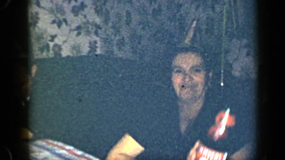 1948 New York home-movie frame of a woman seated indoors at a party table with a beverage bottle in front of her.