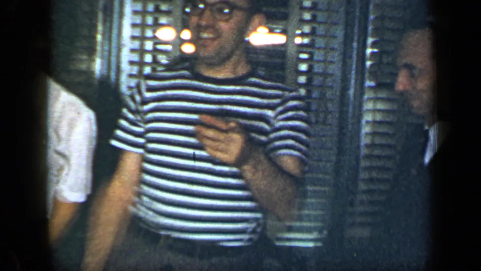 1948 New York party frame with a man in a striped shirt gesturing beside other adult guests in a private home interior.