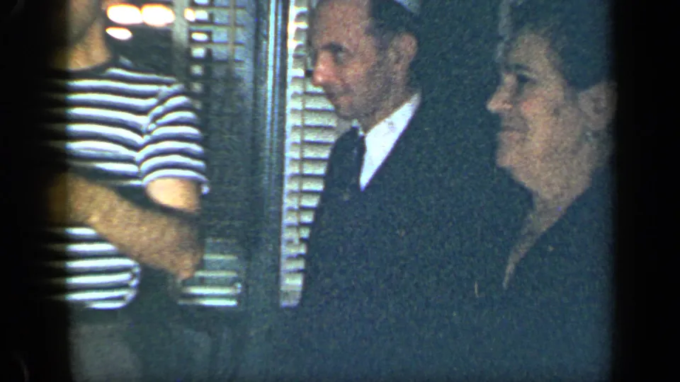 1948 New York home-party frame showing several adult guests, including a suited man and a woman in dark clothing, near interior shutters.
