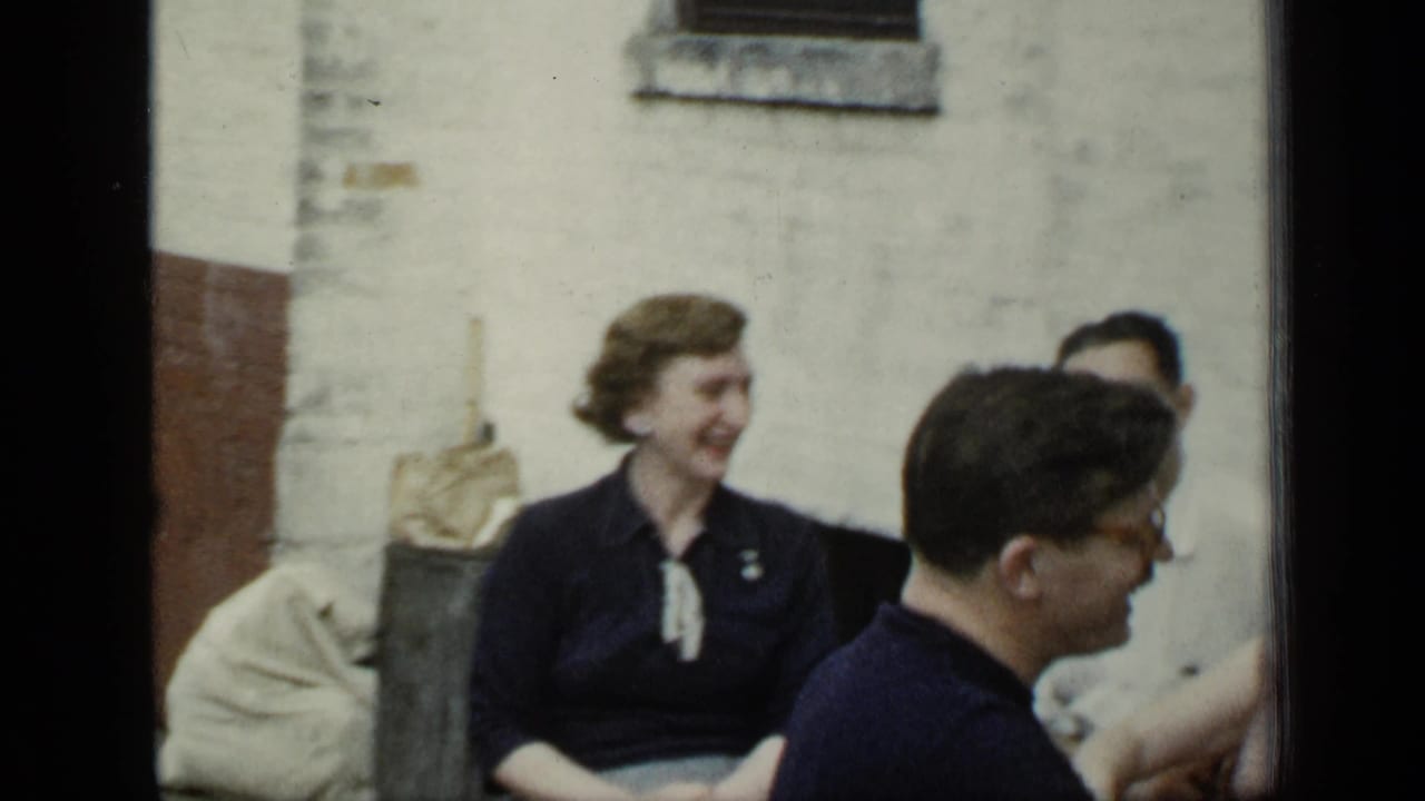 Adults seated outdoors near a pale wall as a man in glasses turns toward others.