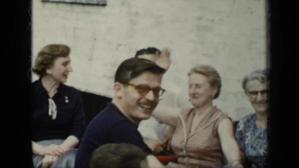 1948 New York outdoor gathering with a smiling man in glasses, seated women, and one raised hand near a pale wall.