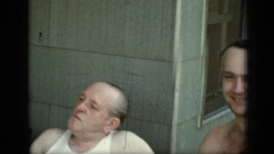 1948 New York home-movie frame with a seated man in a white tank top and another smiling adult near a porch or patio wall.