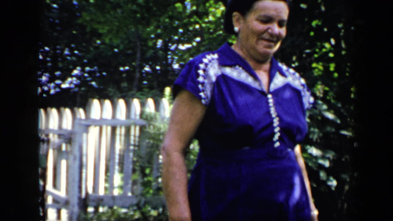 Adult woman in a purple dress walks beside a white picket fence in a leafy garden.