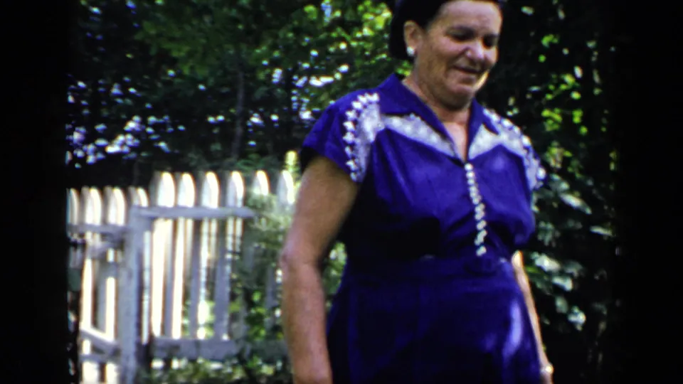 In a 1948 New York 8mm home-movie frame, an adult woman in a purple dress with white trim walks through a sunlit garden near a white fence.