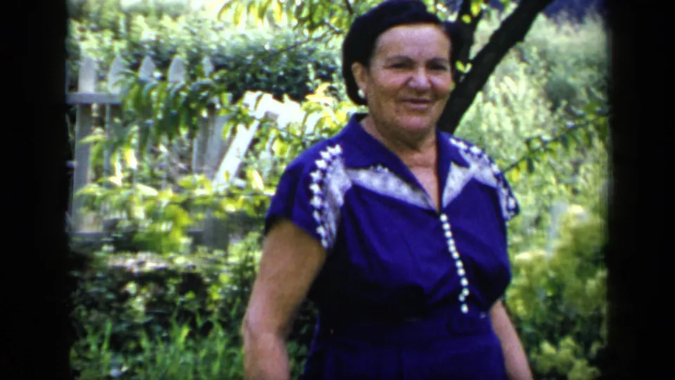 A 1948 New York home-movie frame shows an adult woman in a purple dress moving through a green garden with trees and bright foliage behind her.
