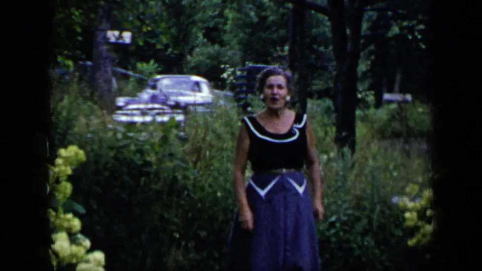 Adult woman in a dark sleeveless top and blue skirt walks toward the camera through a wooded garden, with a parked car behind her.