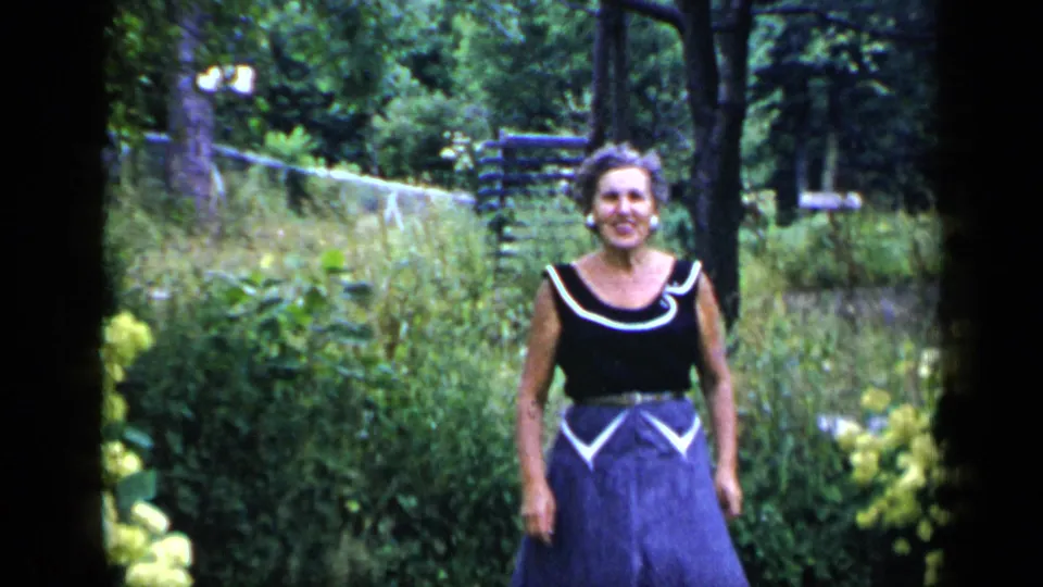 Adult woman pauses in a wooded New York garden area, wearing a dark top and blue skirt with white trim, consistent with 1948 metadata.