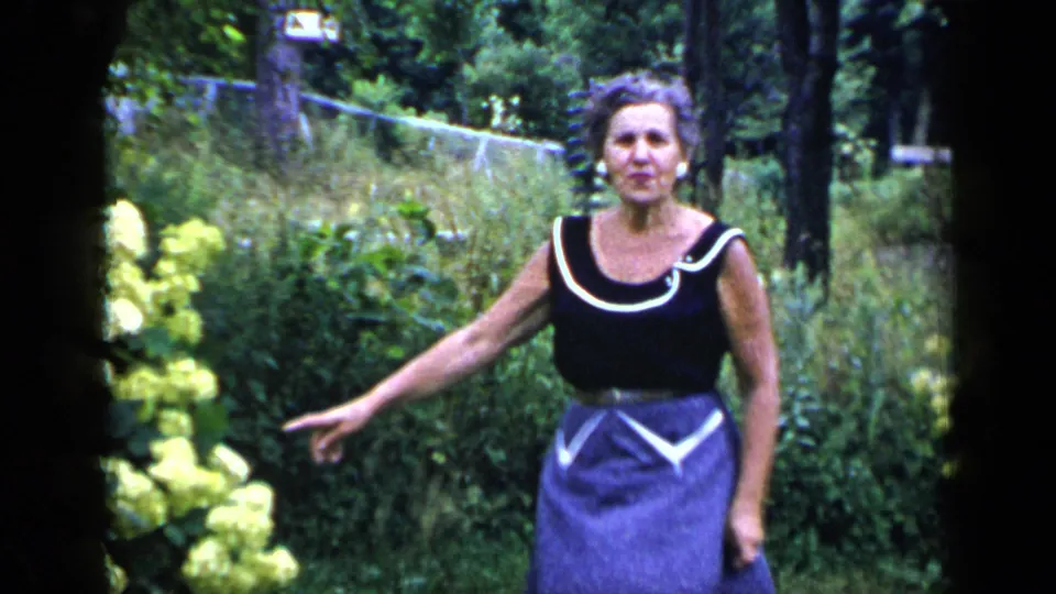 The woman stands beside yellow hydrangeas and points across the garden in this 1948 New York home-movie frame.